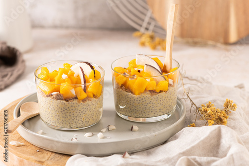 Two glasses of creamy chia seed pudding topped with vibrant diced mango and fresh coconut slices served on a ceramic plate with wooden spoons for a healthy breakfast or nutritious tropical snack