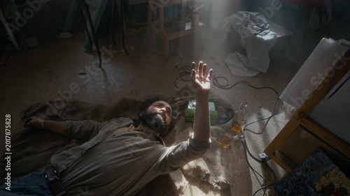 High angle view of tortured male artist lying on dusty floor in his studio, touching sunbeam and drinking whiskey from bottle while suffering from severe depression