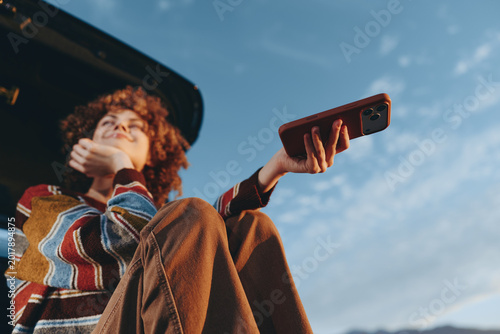 Woman lifestyle outdoors with curly hair smiling while holding smartphone recording, wearing casual striped jacket, standing near car roof, bright sky, sunny backlight, relaxed carefree moment