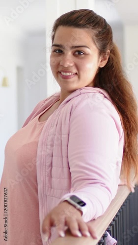 Vertical video: Looking back, woman at home in pink hoodie holding railing, watch on wrist, smiling