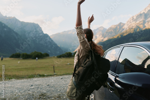 Принти на полотні Young woman with backpack stands beside a car in a mountain valley, arms raised in a joyful, liberated moment after a scenic hike under clear blue skies
