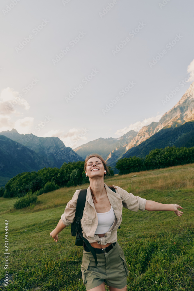 Obraz premium Joyful woman hiking in a green meadow with mountains in the background, wearing a light jacket and backpack. Outdoor adventure and wellness. Embracing nature Outdoors.