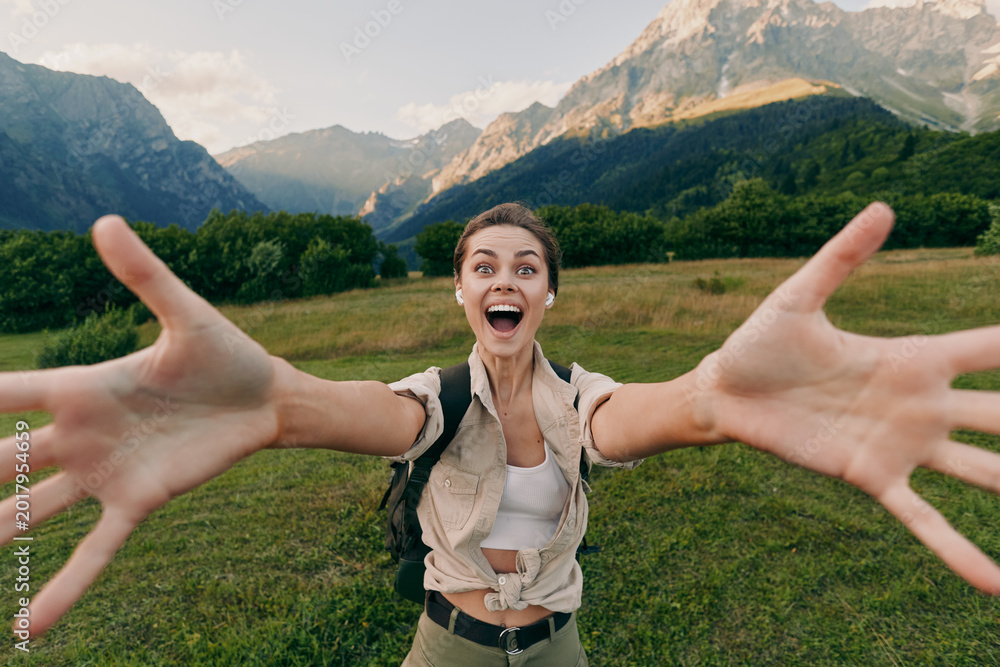 Obraz premium Cheerful woman outdoors, reaching toward the camera with arms wide, sunlit field and mountains behind, casual outfit, joyful lifestyle moment, natural colors