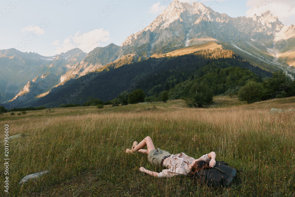 Obraz premium A young woman lies on a sunlit meadow with rugged mountains in the background, resting beside a backpack, conveying relaxation and a sense of outdoor freedom.
