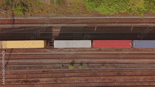 Vertical aerial drone footage looking straight down at cargo freight train transporting colorful shipping containers along railway tracks in the United Kingdom.