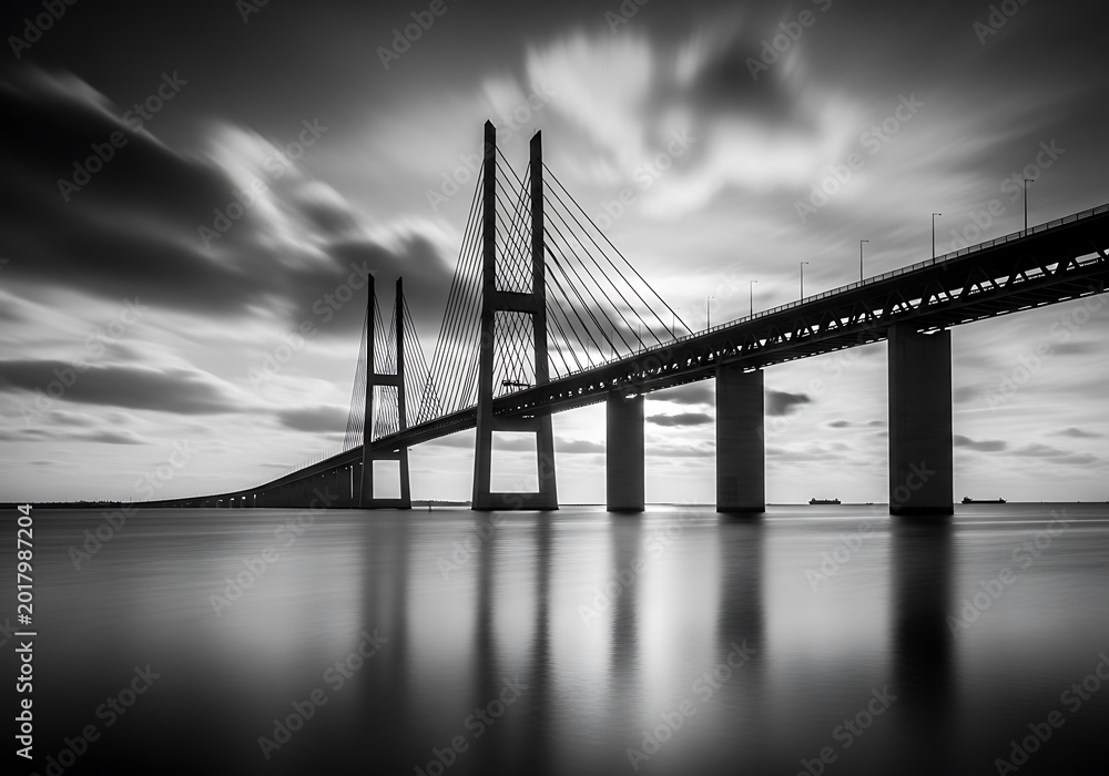 Fototapeta premium Suspension bridge over calm water under cloudy sky black and white