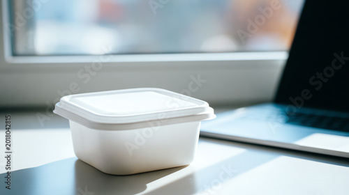 A clean, white takeout container on a desk near a laptop, symbolizing modern dining and convenience in a busy lifestyle.