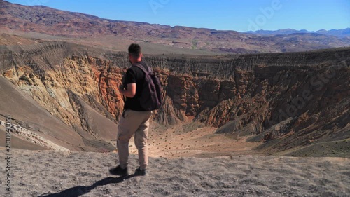 Male hiker with backpack walks along edge of massive Ubehebe volcanic crater in Death Valley National Park, taking in stunning panoramic views of colorful layered rock walls in orange.