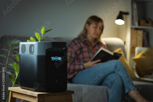 Young woman reading a book on a sofa and using Portable power station to power up lamp during electricity outage. Portable battery as a source of electricity during blackout