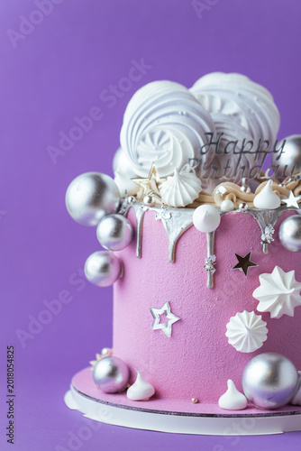 Pink birthday cake with silver drip, meringues, and metallic decorations on purple background. Festive dessert with modern stylish design