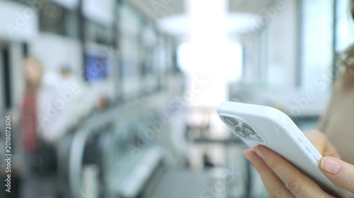 Hand holds smartphone in public transit area. Escalator and people blurred in background. Modern indoor setting with soft lighting. Focus on phone use during commute