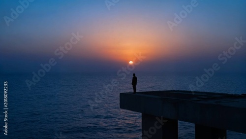 A solitary figure stands in silhouette on the edge of a concrete pier, gazing at a vibrant sunset where the sun hangs low on the horizon, casting warm orange and pink hues across the deep blue sky and