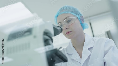 A scientist in a lab coat looks closely at samples through a microscope. The person wears safety goggles and gloves. This takes place in a modern laboratory setting.