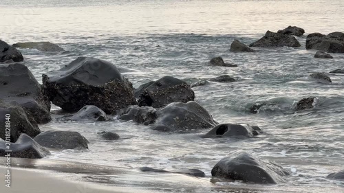 Gentle Ocean Waves Caress Dark Rocks on a Tranquil Sandy Beach at Golden Hour