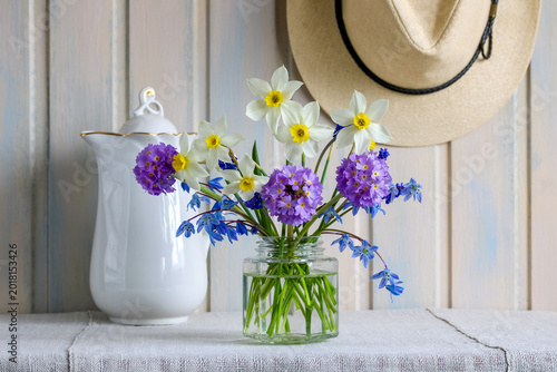 Rustic spring still life with fresh narcissus flowers, primrose bouquet and blue scilla in glass jar beside white pitcher and straw hat