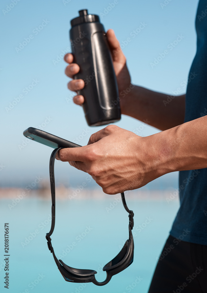 Fototapeta premium Bottle, swimmer and hands of man with phone outdoor for texting, networking or contact at practice. Typing, fitness and athlete with cellphone for social media on mobile app at competition training.