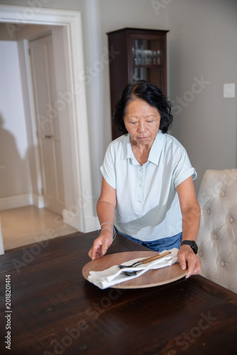 Senior Chinese woman arranging charger with napkin cutlery at table in blue shirt with smartwatch