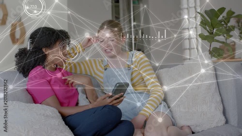 Two teenage females tapping smartphone on sofa, generating animated HUD charts to share tech data
