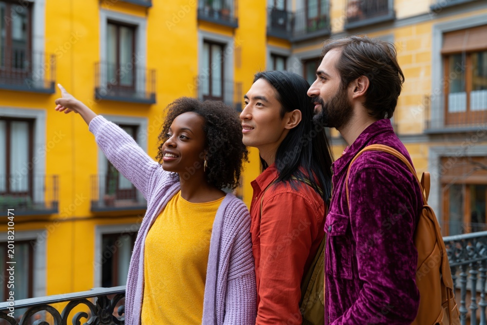 Obraz premium Trio of friends standing on a balcony in a colorful city pointing at buildings for travel banner background and lifestyle imagery
