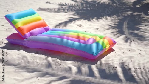 A rainbow-colored inflatable float rests on a sandy beach with palm tree shadows