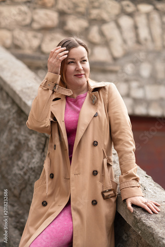 Slika na platnu Trenchcoat Woman Fashion: Woman poses outdoors in beige trench coat, day, adjusting hair