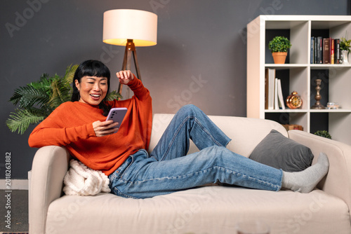 Happy Asian woman lying on sofa and using smartphone in living room, Leisure and Technology concept. Relaxed mature lady browsing social media or watching video on mobile phone at home. Lifestyle.