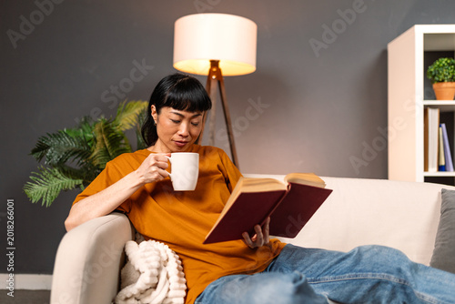 Asian woman reading book and drinking coffee on sofa at home, Leisure and Lifestyle concept. Mature female relaxing with literature and hot beverage in cozy living room. Weekend domestic hobby concept