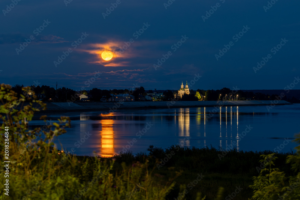 Fototapeta premium A beautiful night landscape with a full moon rising over a river. Moonlit path on the water. The Church of St. Simeon the Stylite is visible in the distance. Veliky Ustyug, Vologda Oblast, Russia.