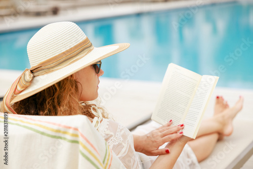 Obraz na plátne Woman reading book by poolside relaxing during summer vacation