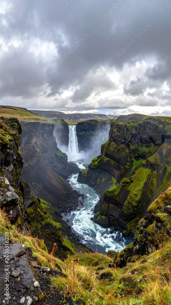 Obraz premium Majestic waterfall cascades into a deep, moss-covered canyon under dramatic, cloudy skies