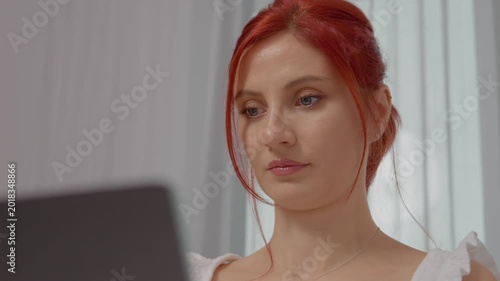 A woman with red hair is focused on her laptop, typing away and occasionally glancing at the screen. The setting is bright and inviting, enhancing her concentration.