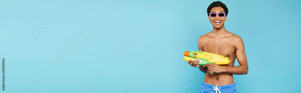 Naklejka premium Handsome young man enjoying a vibrant summer day with a water gun by a bright blue backdrop