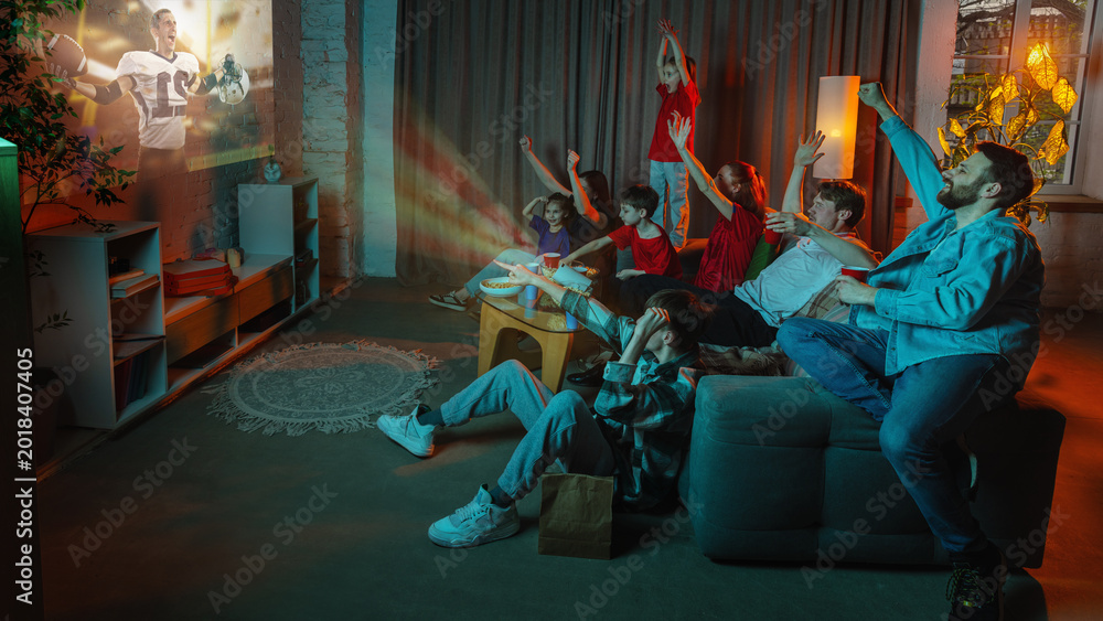 Fototapeta premium Big family watching American football projection at home with cheering children. Concept of live sports streaming, fan night, subscription media, home cinema, family fun, and weekend viewing.
