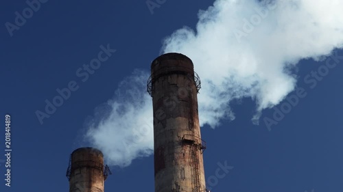 Close up of industrial smokestacks with white smoke emissions against dramatic blue sky. Handheld static 4K footage of industrial pollution and heavy industry infrastructure.