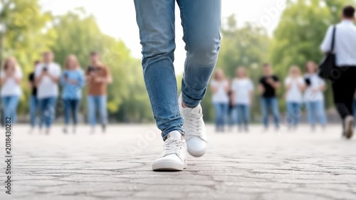 Tracking shot of person walking toward camera on paved park walkway with blurred pedestrians and trees in background during daytime spring