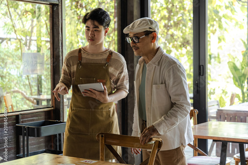 Small business cafe owner guiding young barista, father and son discussing table setup while holding tablet, small business workflow planning and service improvement in cozy restaurant and coffee shop