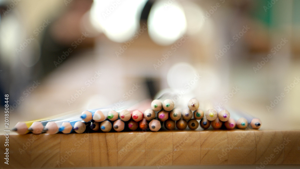 Fototapeta premium Colored pencils arranged on a wooden table with soft blurred classroom background