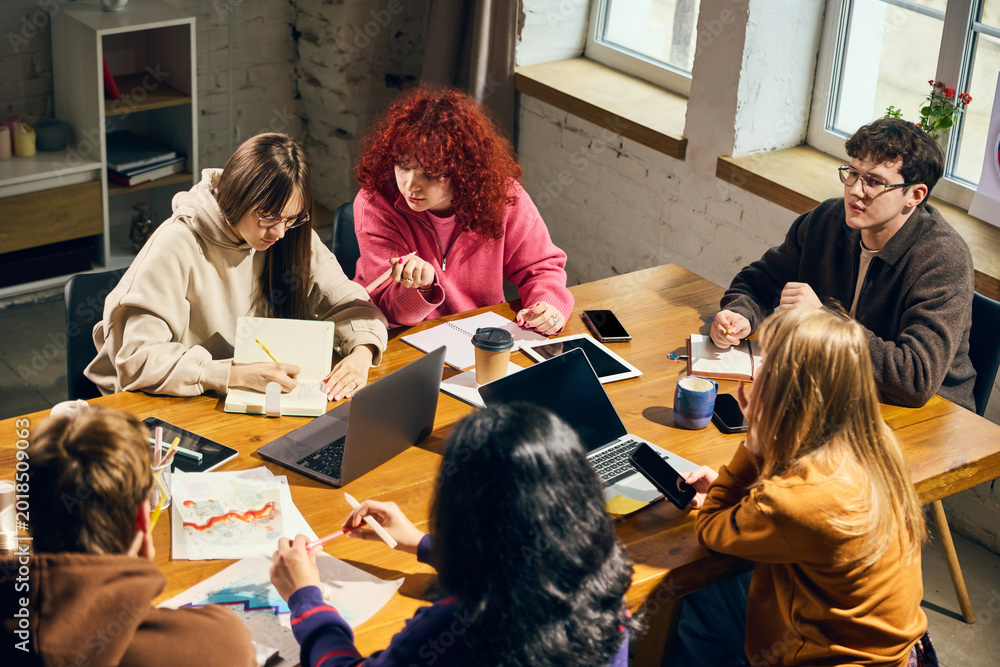 Obraz premium Young people collaborating around wooden table with laptops and charts. Concept of business planning, creative agency work, student workshop, teamwork, analytics, and office training.