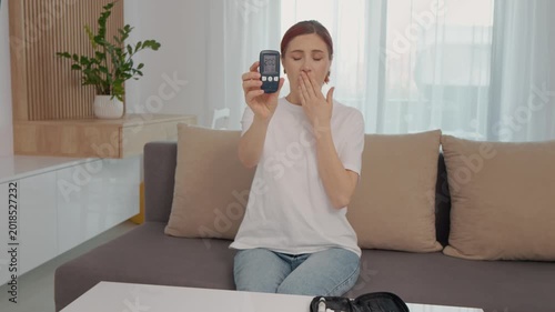 A woman shows her glucometer reading of 200 mg dL while sitting on a couch at home. This shows her concern about high blood sugar levels and diabetes.