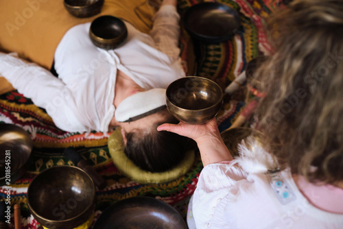 High angle view of a therapist playing a Tibetan singing bowl near a woman resting with an eye pillow. Private sound bath rituals support stress relief, deep relaxation, and emotional balance.