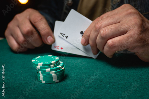 A man sits at a green table with poker chips stacked in front of him. He holds two playing cards with aces on them. The action takes place in a night casino