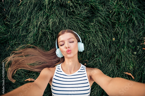 Schilderij op canvas Young woman lying on grass wearing headphones taking a playful selfie with pucke