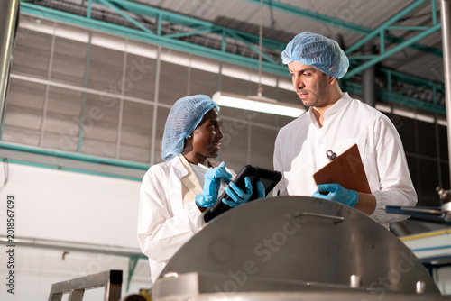 Diverse factory team workers inspecting large metal tank, Caucasian man African American woman using digital tablet checking manufacturing, food processing plant production line machinery.