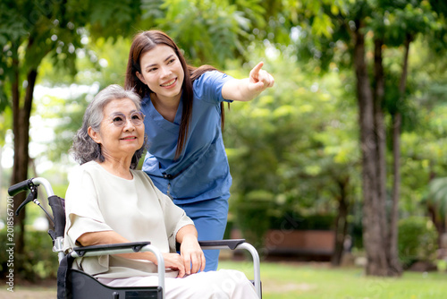 Caucasian female caregiver encouraging senior patient on wheelchair in garden, professional nurse support elderly woman for rehabilitation and mental health therapy outdoors, medical and assistance.