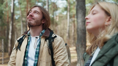 Peaceful hikers resting among pine trees. Caucasian man smiling while breathing fresh forest air beside woman. Travel partners enjoying calm outdoor atmosphere during countryside adventure.