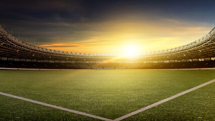 A wide-angle view of a professional football stadium at sunset. The empty arena features a lush green field under a dramatic golden hour sky with a bright lens flare