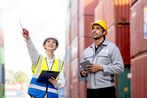 Workers with safety vest and helmet discussing at logistic shipping cargo container yard. Male engineer with tablet works together with woman supervisor holds worky talky and clipboard during inspects