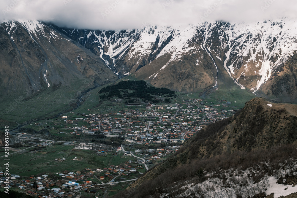 Obraz premium Panoramic view of Stepantsminda village nestled in the Caucasus Mountains near the Russian border, Georgia. Alpine settlement under snowy peaks and low clouds in the Kazbegi region