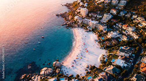 Aerial view of a bustling beach with white sand and turquoise water, lined with buildings and lush green trees.