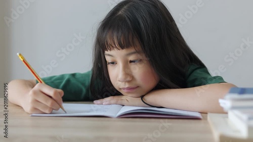 Close up selective focus of demotivated Asian kid girl resting her head on a desk in daylight. She looks overburdened by too much homework, illustrating academic pressure and childhood study burnout.